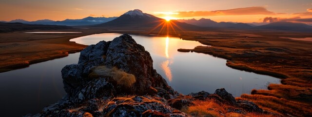  The sun sets over a mountain, with a body of water in the foreground and mountains in the background