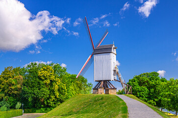 Old windmill on green hill in Brugge city, blue sky white clouds background in summer sunny day, typical wooden wind mill with sails and path in park with green trees, West Flanders province, Belgium