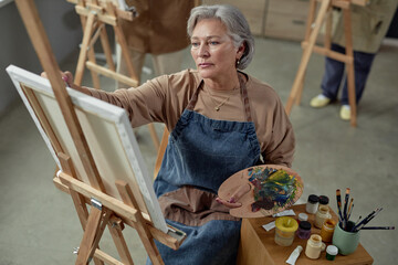 High angle portrait of creative mature woman painting picture on easel in art studio and holding palette pursuing inspiration