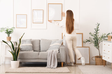 Poster - Young woman hanging blank frames on white wall in light living room