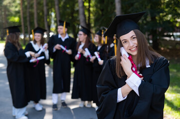 Wall Mural - Group of happy students in graduation gowns outdoors. A young girl boasts of her diploma.