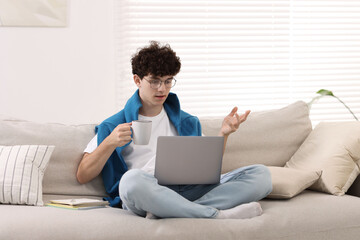 Wall Mural - Teenager holding cup of drink working with laptop on sofa at home. Remote job