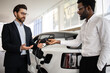 © sofiko14 - Dealer explains electric SUV charger to businessman in car dealership showroom. Professional interaction between car dealer and customer with new electric vehicle in background.