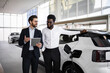 © sofiko14 - Dealer and businessman discuss electric SUV inside modern car dealership. Business context with focus on eco-friendly transportation. Conversation and decision-making process.