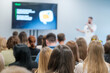 © Anton Gvozdikov - Diverse audience in a business conference listening to a speaker with a presentation displayed on a screen.