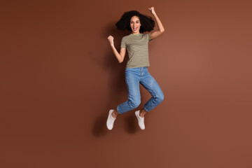 Wall Mural - Full length photo of adorable lucky girl wear khaki t-shirt jumping high rising fists empty space isolated brown color background