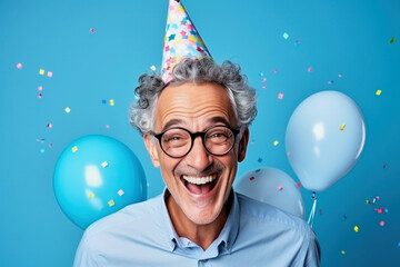 Wall Mural - Happy excited amused birthday man wearing festive cap with colorful confetti and helium balloons on a blue background