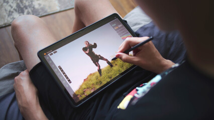 Fototapete - Boy with digital stylus and tablet in hands sits in cozy room and draws 3d model. Teenager creates new skin using 3d modeling program. Young game designer remote studying. Game development. Close up.