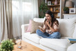 © Srdjan - Simple living. Young beautiful relaxed woman sitting alone at home on sofa, listening podcast or music on her wireless headphones connected to her smart phone. Freedom concept and weekend activities.