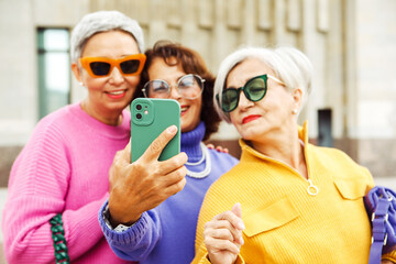 Three senior women taking selfie photo together outdoors, having fun and smiling while wearing colorful and sunglasses.