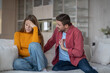 © DimaBerlin - Squabble of married couple on sofa in living room. Abusive man making excuses aggressively attacks upset woman covering face with hand holding back tears. Disgruntled irritated guy offended girl fight