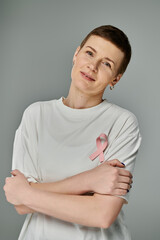 A woman with short brown hair, wearing a white t-shirt with a pink ribbon, stands with her arms crossed in front of a gray background.