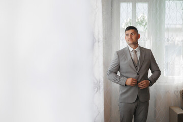 Wall Mural - A man in a suit is standing in front of a window, adjusting his tie. Concept of formality and professionalism, as the man is dressed in a suit and tie