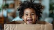 © familymedia - A joyful and smiling toddler with curly hair is sitting in a cardboard box inside a home. The setting is filled with warm tones and the child looks very happy and playful.
