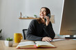 © LIGHTFIELD STUDIOS - A young woman in a grey sweater smiles while talking on the phone at her desk, with a notebook and coffee cup in front of her.