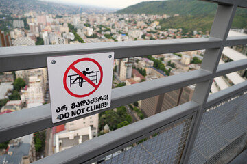 A red warning sign on a rooftop saying DO NOT CLIMB in English and Georgian, is attached to a metal railing with blurry city buildings in the background.