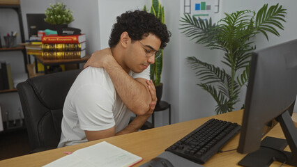 Canvas Print - Young man with back pain sitting in an office, surrounded by books and plants, holding his shoulder in discomfort while working at a computer.