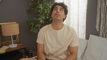 Sticker - Young hispanic man sitting on a bed in a cozy bedroom, looking up thoughtfully, surrounded by home decor and indoor plants, suggesting a relaxed and introspective moment at home.