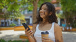 © Krakenimages.com - Brunette hispanic woman in an urban park holding a coffee cup and smartphone outdoors with trees and buildings in the background.