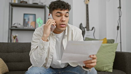 Wall Mural - A handsome young man in a casual outfit attentively on a phone call while holding a document in a modern living room.