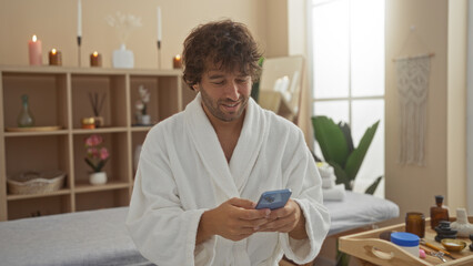 Wall Mural - Handsome young man in a spa wearing a robe checking his phone while surrounded by wellness decor and candles, creating a serene interior scene in a beauty salon.