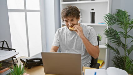 Poster - A bearded man snacking while working on a laptop in a modern home office setting