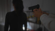 © Krakenimages.com - An armed person points a handgun at a woman's back in a dimly lit indoor setting, implying a tense confrontation.