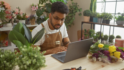 African american man working on laptop in a flower shop surrounded by fresh blossoms and greenery.