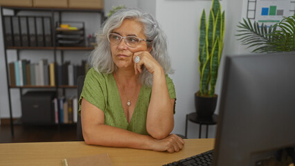 Poster - Elderly woman with grey hair and glasses sitting thoughtfully at a desk in an office room with bookshelves and plants in the background.