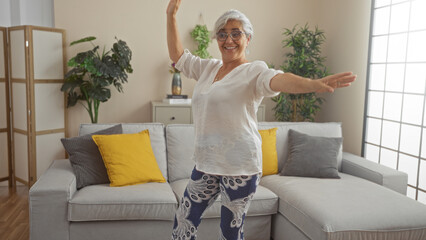 Poster - Elderly woman dancing joyfully in a cozy living room with yellow and gray pillows on the sofa, surrounded by indoor plants and soft natural light