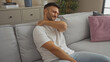 © Krakenimages.com - A handsome young hispanic man with a beard in an apartment living room clutching his sore shoulder while sitting on a comfortable sofa.