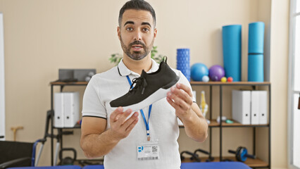 Sticker - A bearded hispanic male therapist exhibits a sports shoe in a rehabilitation clinic's therapy room.