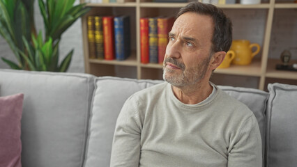 Mature bearded man sitting thoughtfully on a gray couch in a cozy living room interior with bookshelf
