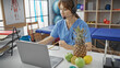 © Krakenimages.com - A focused woman in scrubs uses a laptop in a physiotherapy clinic, adorned with anatomy posters and exercise equipment.