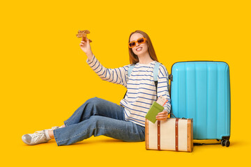 Sticker - Smiling young woman with her luggage sitting on floor, holding passport and  airplane on yellow background. Travel concept