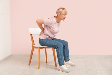 Poster - Mature woman suffering from back pain while sitting in chair on pink background