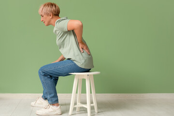 Poster - Mature woman suffering from back pain while sitting on chair on green background