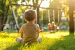 © InfiniteStudio - Happy and joyful three-year-old boy sitting on the grass in a park, children playing on swings in the background, sunny day.