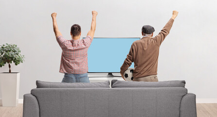 Wall Mural - Rear view shot of a father and son watching football and cheering in front of tv