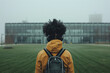 © yaqui_villegas - A person wearing headphones and a backpack walks across a grassy field towards a modern glass-fronted building, shrouded in a misty, fog-covered atmosphere, symbolizing solitude and reflection.