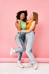 Wall Mural - Two women in casual attire embrace, smiling against a pink backdrop.