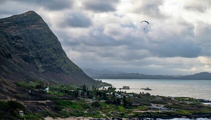 Canvas Print - Scenic view of a coastal landscape with a mountain, cloudy sky, and a paraglider over the ocean