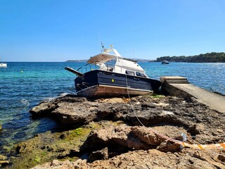 Canvas Print - Beached boat on a rocky shore with a clear blue sky in the background. Palma Nova, Mallorca, Spain