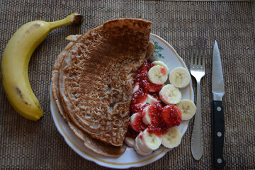 Wall Mural - Fresh sliced bananas with sweet raspberry jam and chocolate pancakes arranged in a light clay plate, a yellow whole banana, a kitchen knife and a fork.