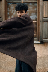 Wall Mural - Fashionable young man in brown coat and scarf standing in front of window with arms outstretched