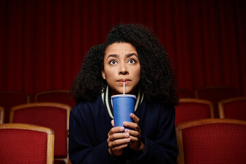 A young woman in casual clothes sits in a theater seat, holding a cup and staring up in surprise, seemingly engrossed in a movie.