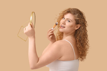 Wall Mural - Beautiful young woman with foundation on her face holding mirror and makeup brush against beige background