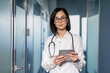 © Liubomir - Confident female doctor with glasses and stethoscope holding digital tablet in modern hospital hallway. Professional healthcare worker with technology, ready to assist patients.
