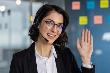 Smiling businesswoman wearing headset waving hand in modern office setting. Professional attire and friendly demeanor suggest customer support, communication, and positivity.