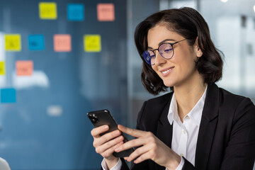 Poster - Confident businesswoman wearing glasses holding smartphone in modern office. Thoughtful professional checking messages on phone surrounded by sticky notes. Evening working atmosphere